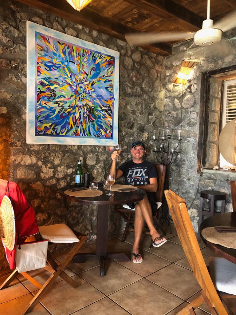 Francesco raising a glass of wine in a stone-walled Caribbean restaurant with a colorful abstract painting