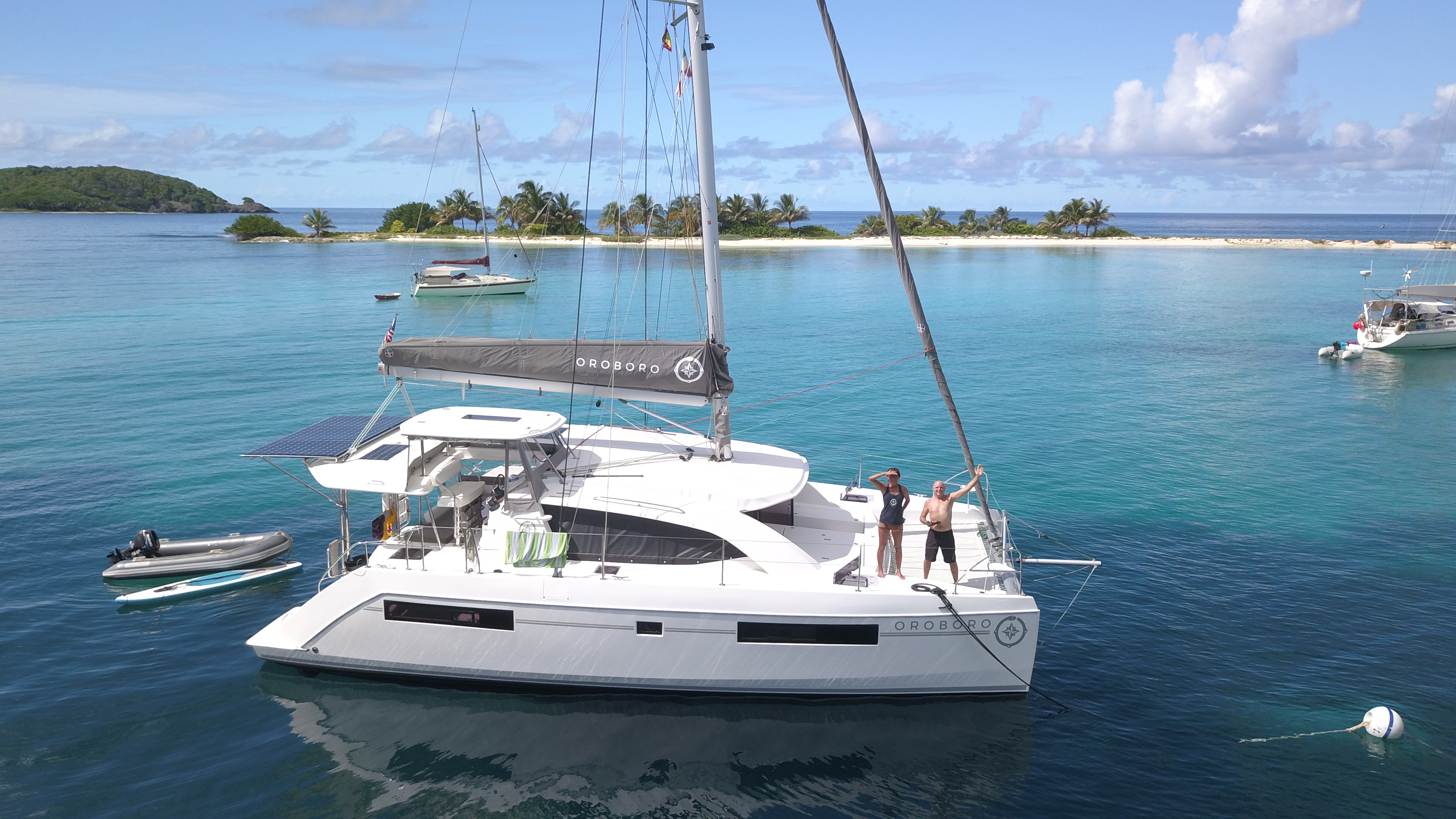 Oroboro at anchor off Carriacou — drone view with two people waving from the deck