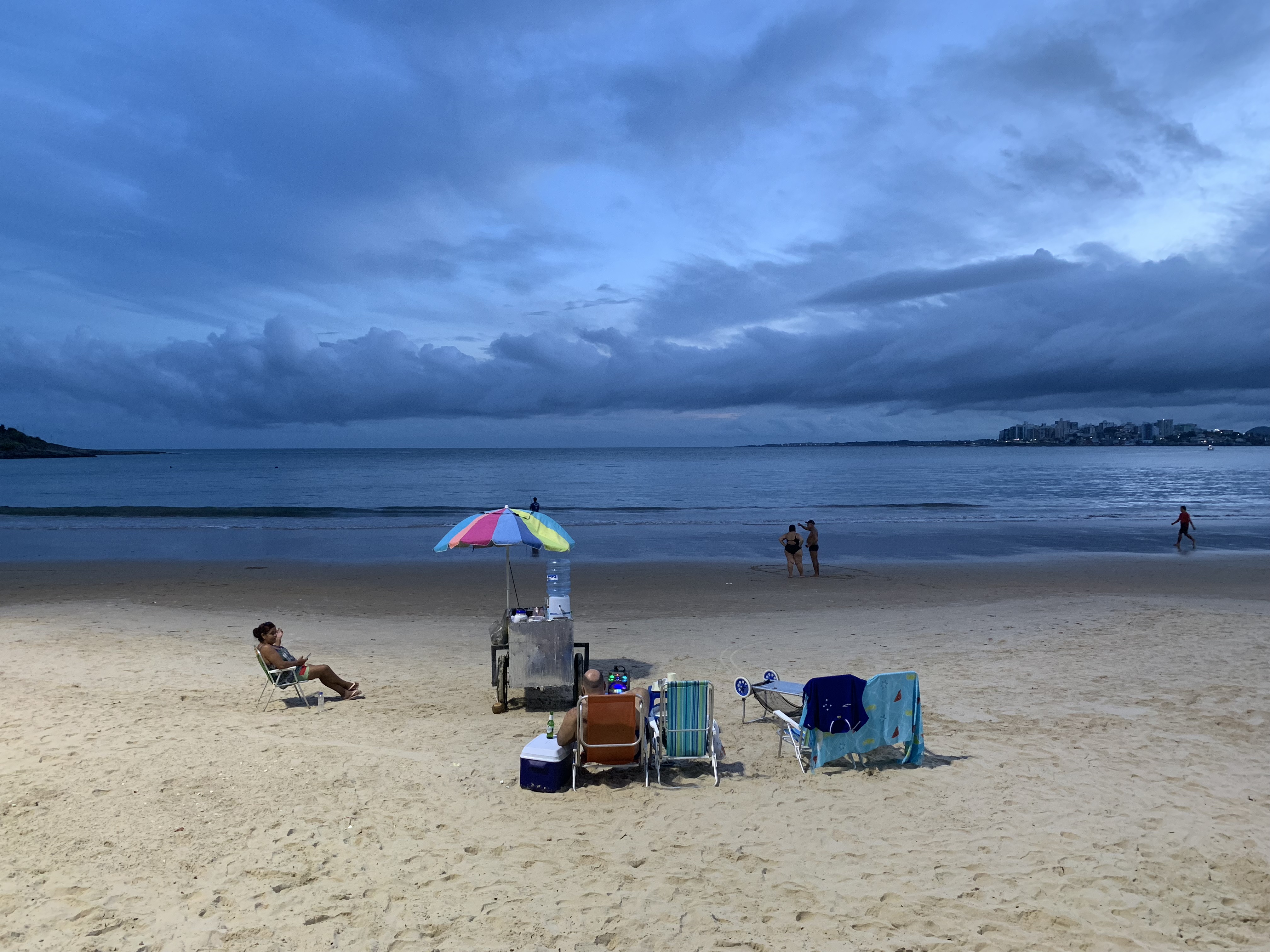 Tarde de sábado na praia — moradores se acomodam ao redor do carrinho de um ambulante enquanto o entardecer tempestuoso avança pelo mar