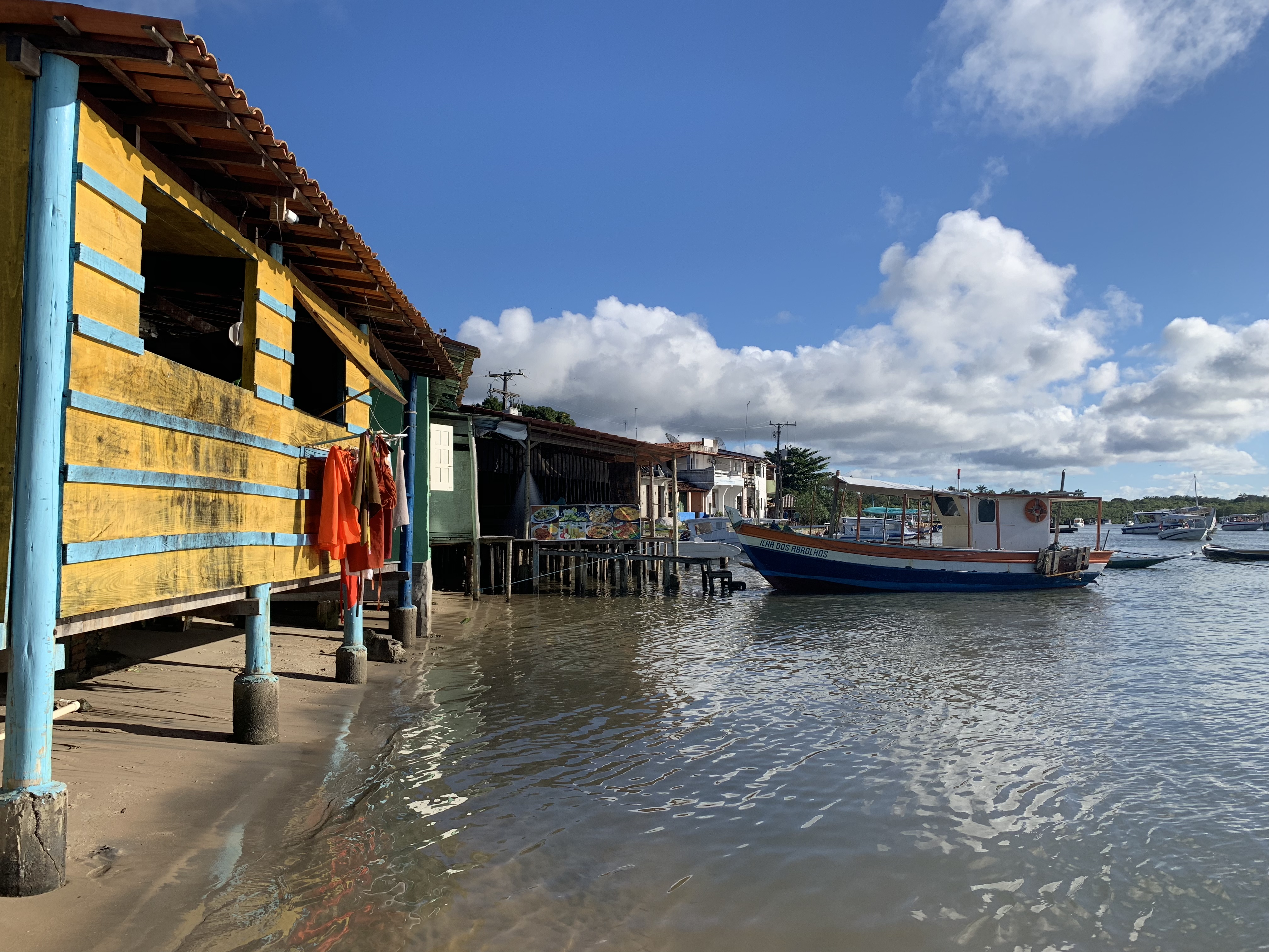 Casas coloridas amarelas e azuis sobre palafitas na orla da vila de Boipeba, com um barco de pesca ao lado