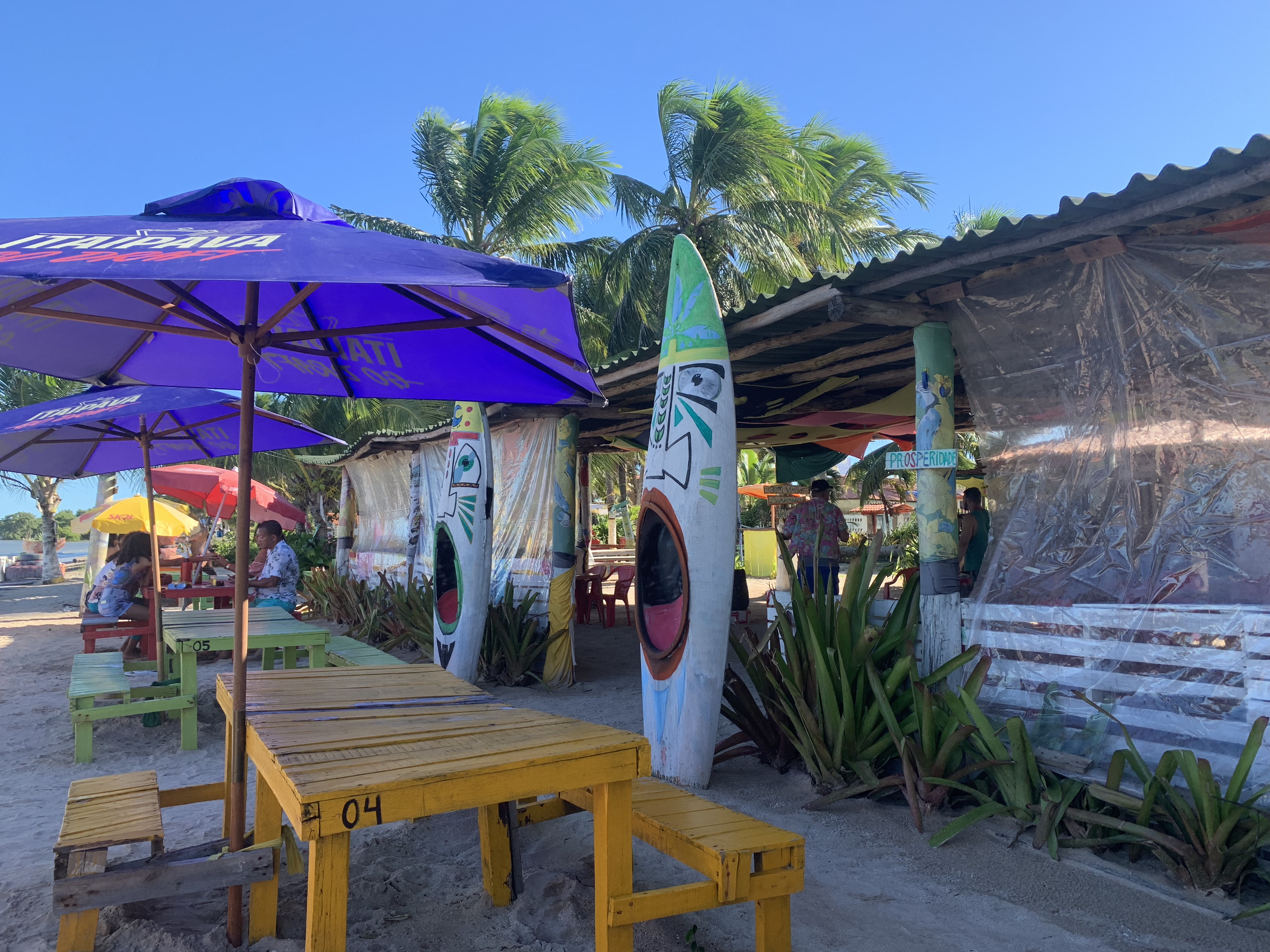 Um colorido bar de praia em Jiribatuba decorado com pranchas de surfe pintadas, mesas de piquenique amarelas na areia