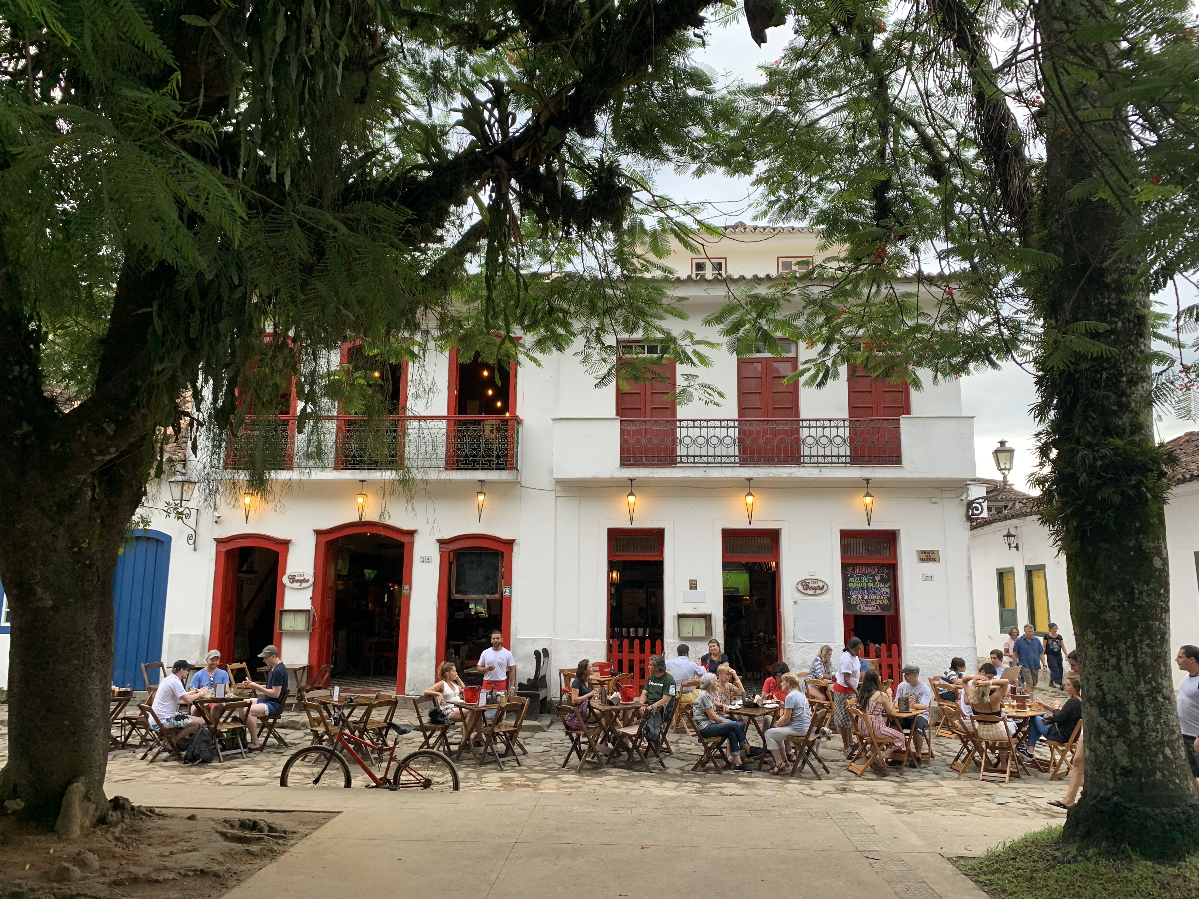 Pessoas jantando em mesas ao ar livre em uma animada praça de Paraty, edificações coloniais com venezianas vermelhas e uma grande árvore ao fundo