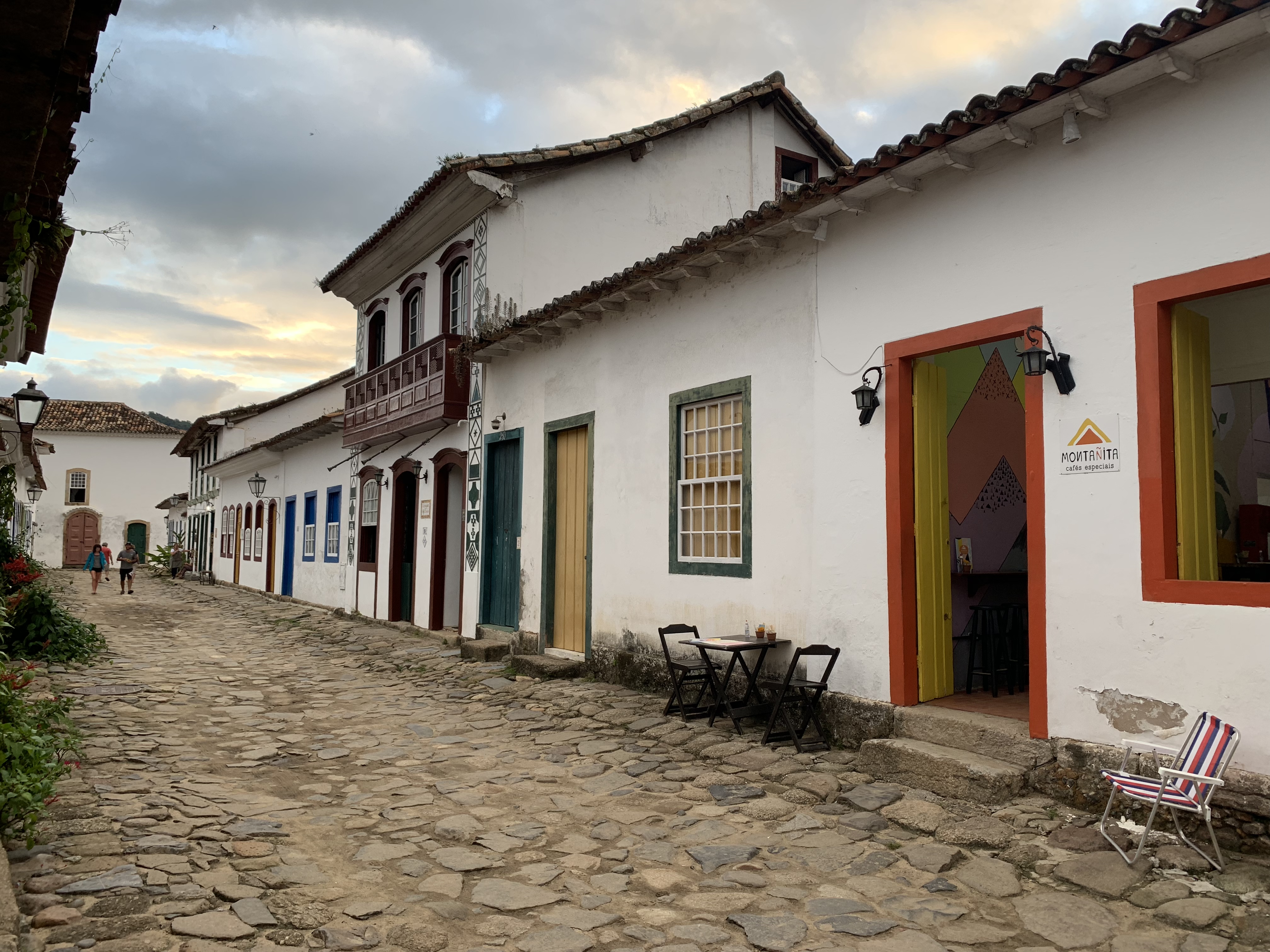 Um café em uma rua de paralelepípedos de Paraty ao entardecer, os edifícios banhados pela última luz quente sob um céu dramático de nuvens