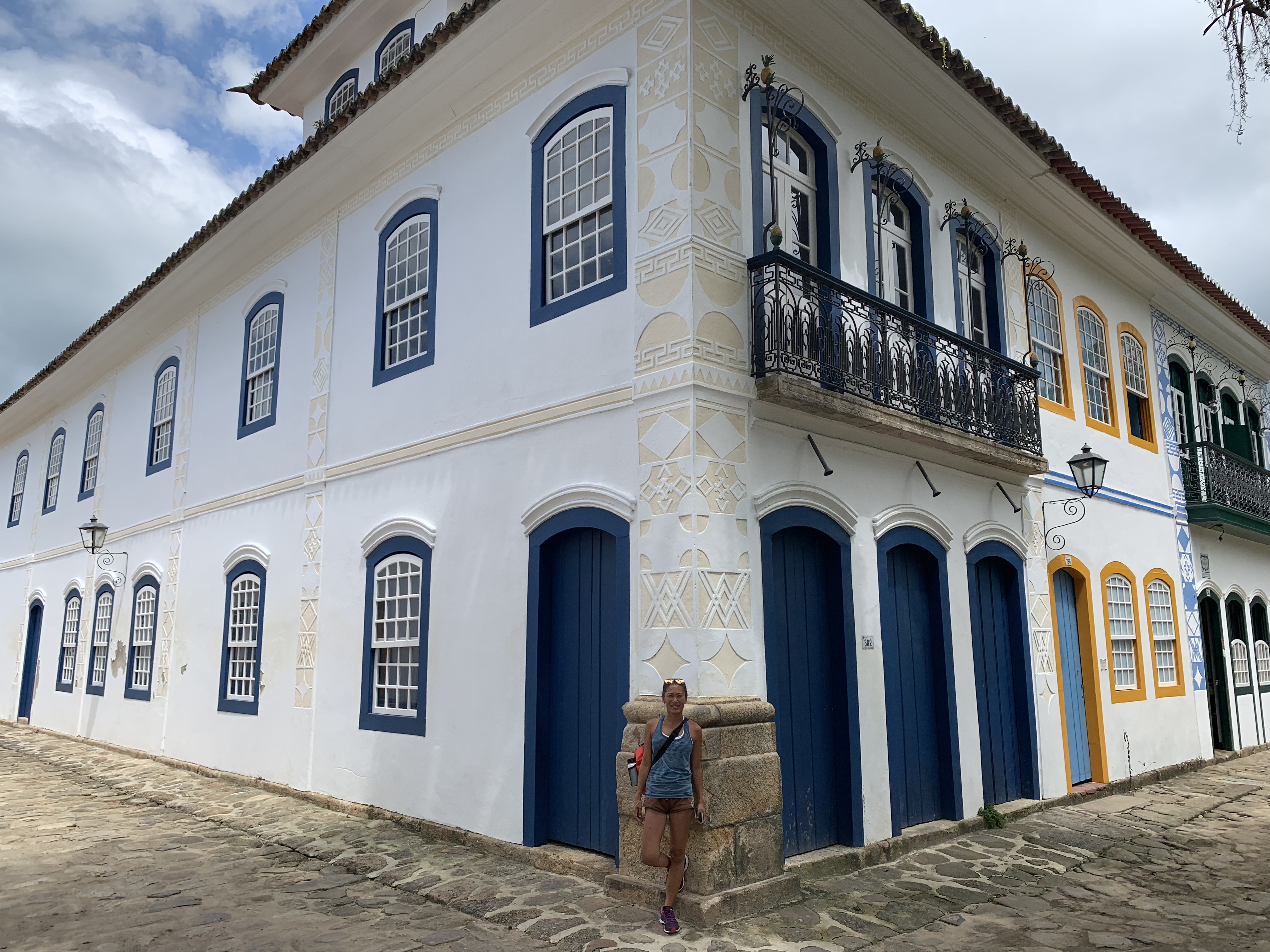 Yuka na esquina de um grande e ornamentado edifício colonial em Paraty com padrões decorativos geométricos na fachada, janelas pintadas de azul e varanda em arco com grades de ferro forjado