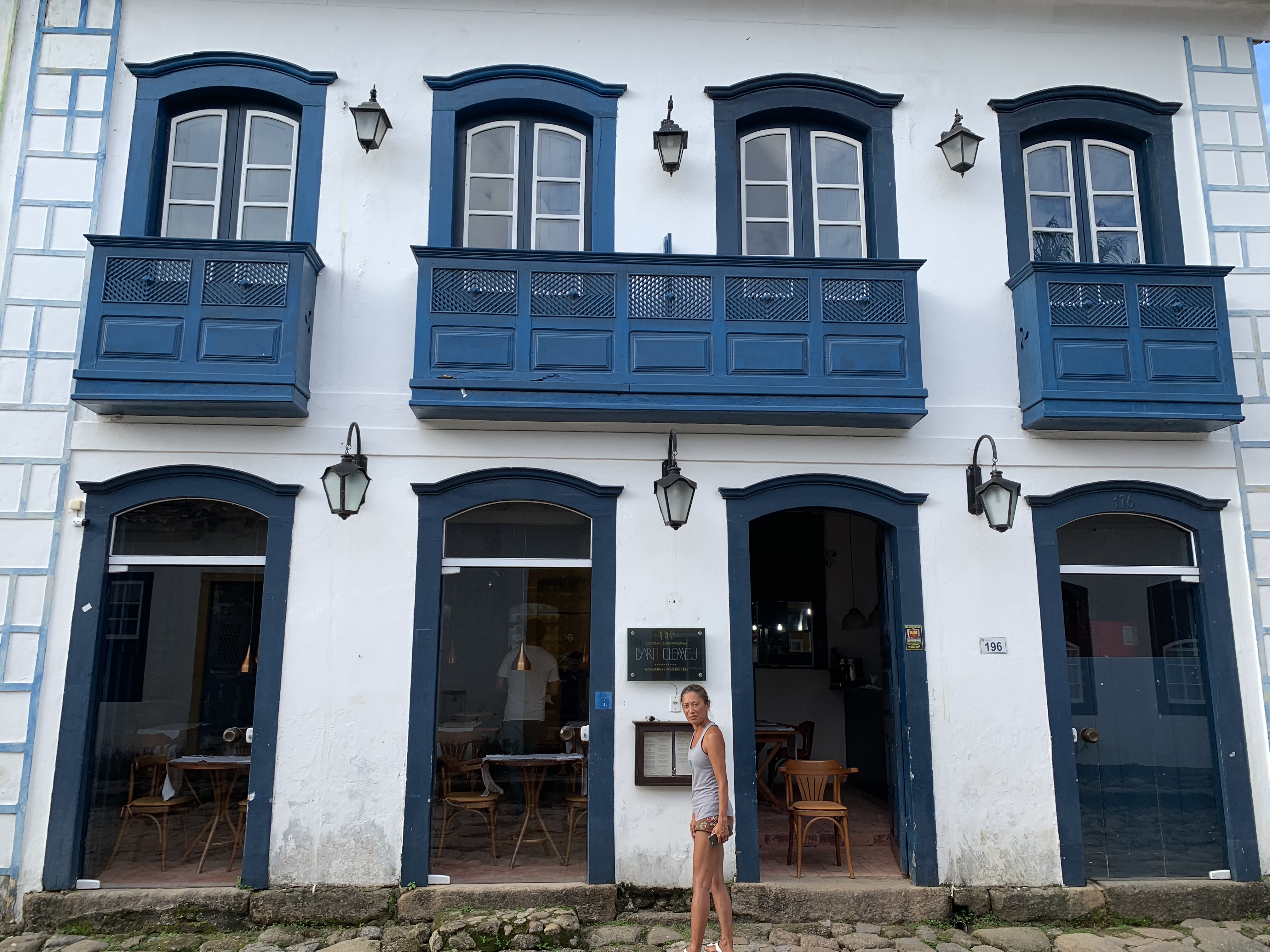 Yuka em frente a um elegante edifício colonial de dois andares em Paraty com varandas em arco azul profundo e portais em arco, lanternas de parede e mesas de café visíveis no interior
