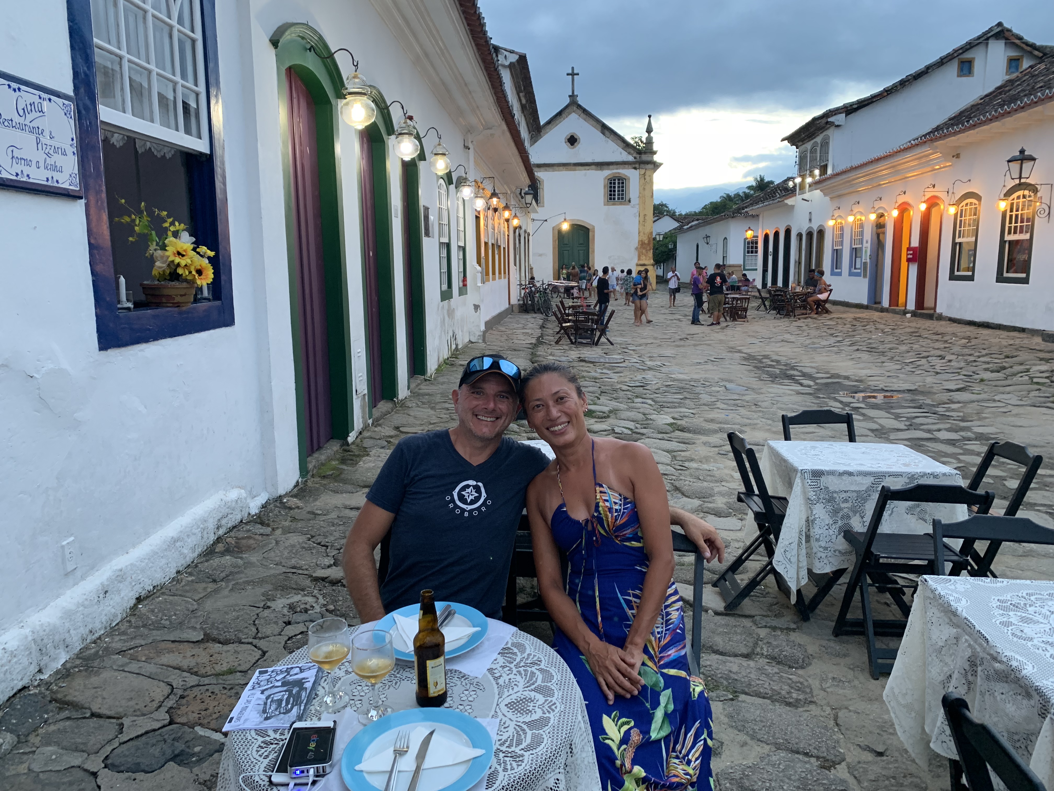 Francesco e Yuka jantando juntos em uma mesa ao ar livre na praça de paralelepípedos de Paraty ao entardecer, com uma igreja colonial e edifícios iluminados visíveis ao fundo