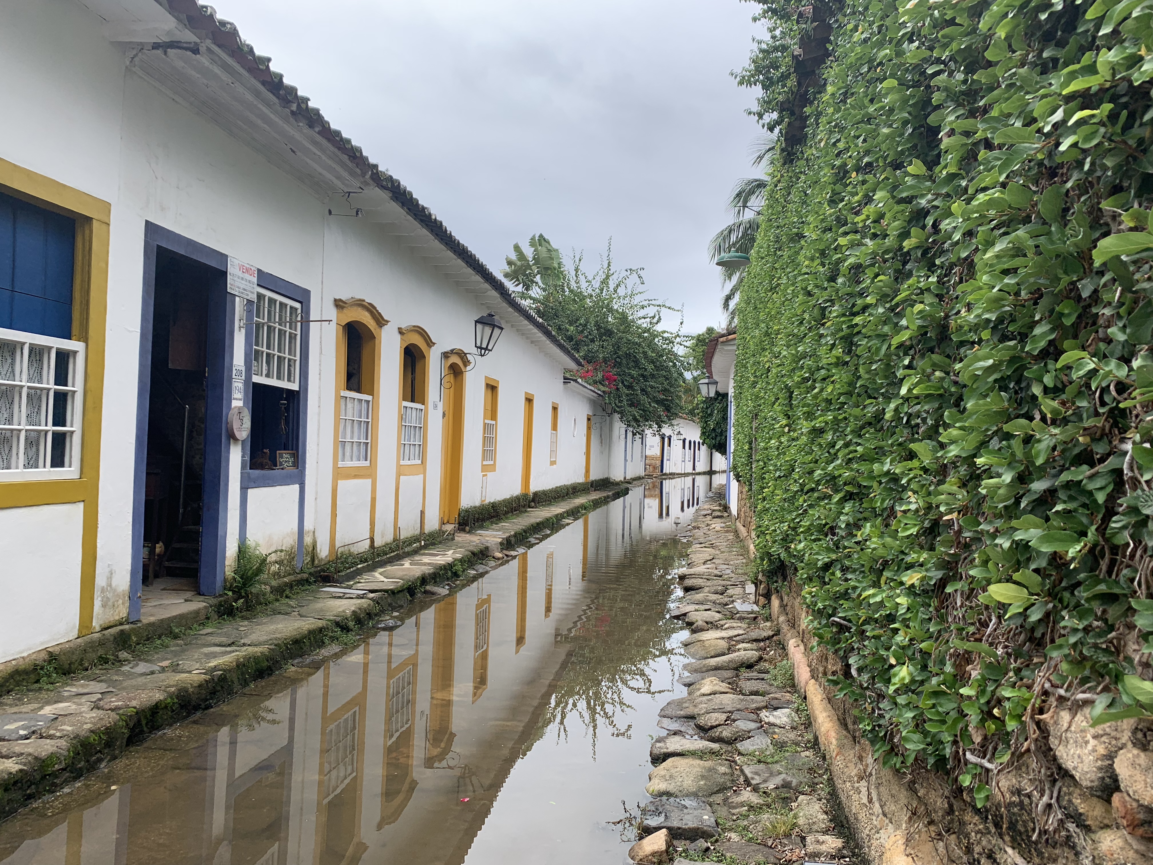 Uma viela estreita em Paraty completamente alagada pela maré alta, a água criando um reflexo perfeito das casas caiadas de branco com janelas de moldura amarela e uma parede coberta de trepadeiras à direita