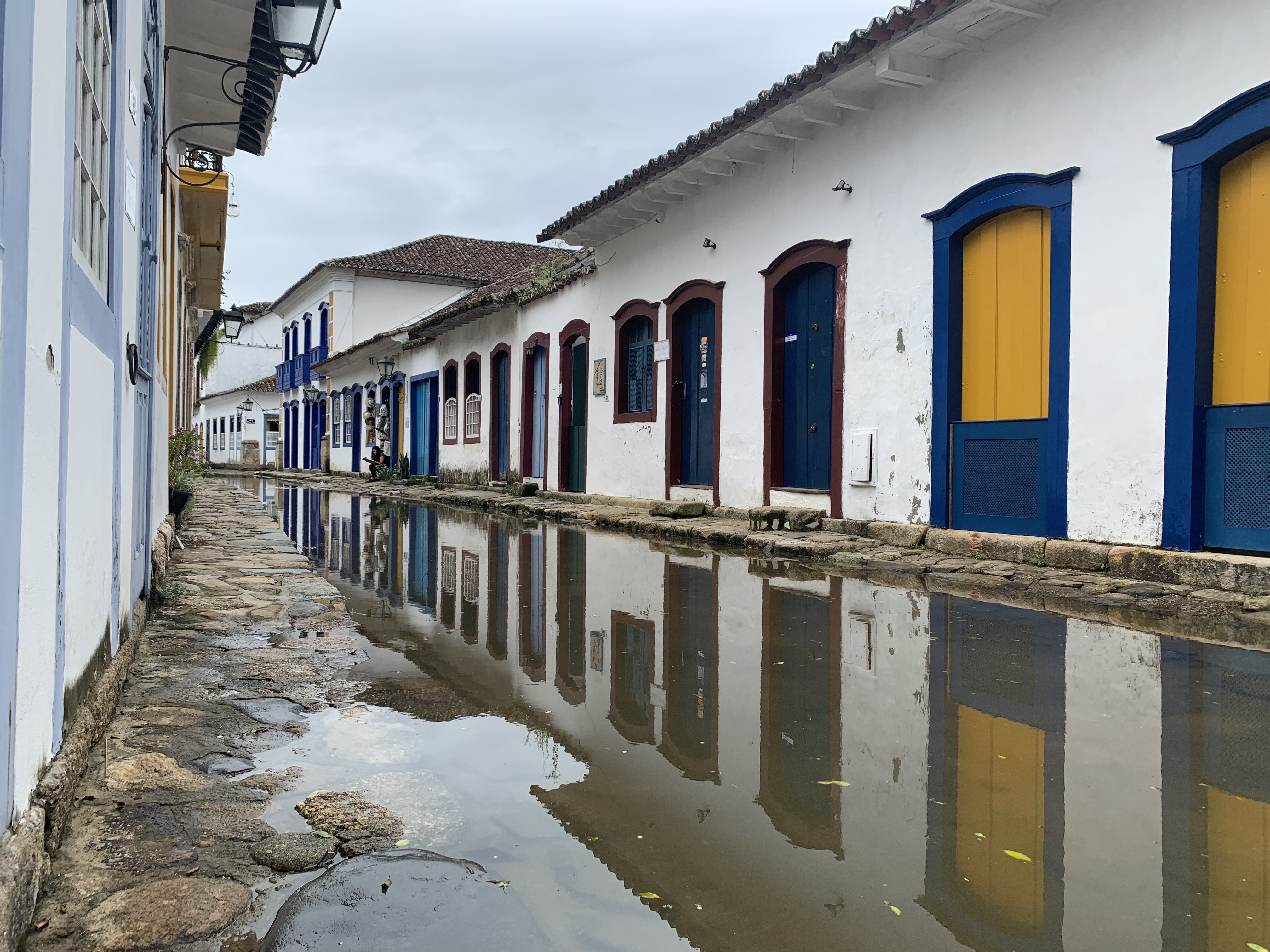 Uma longa rua de Paraty completamente alagada, uma fileira de casas coloniais brancas com portas coloridas em azul, amarelo e verde perfeitamente refletidas na água parada da maré
