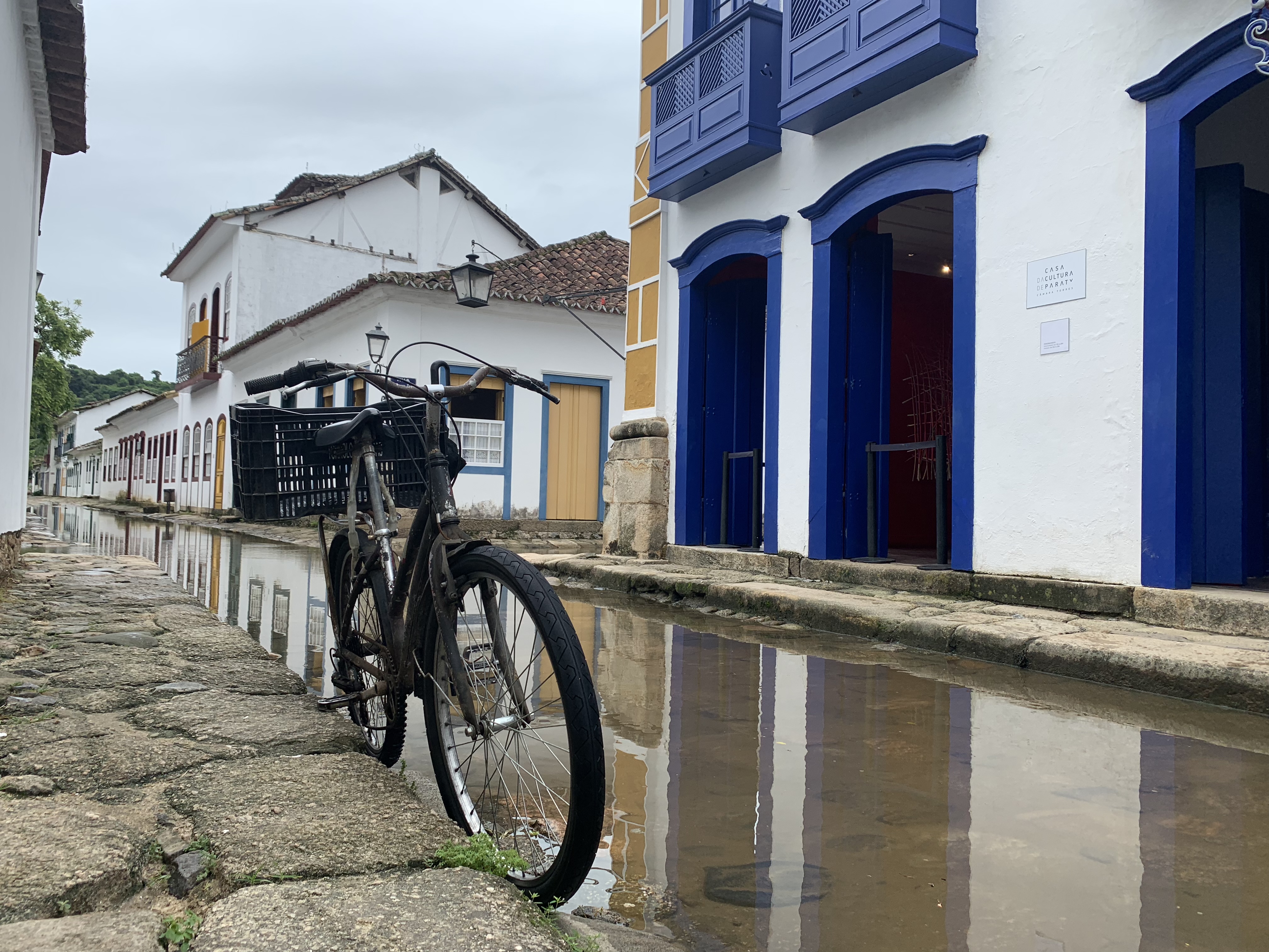 Uma bicicleta preta com cesto estacionada sobre paralelepípedos secos na margem da enchente de maré em Paraty, com edifícios coloniais em azul e ocre refletidos na água parada ao fundo