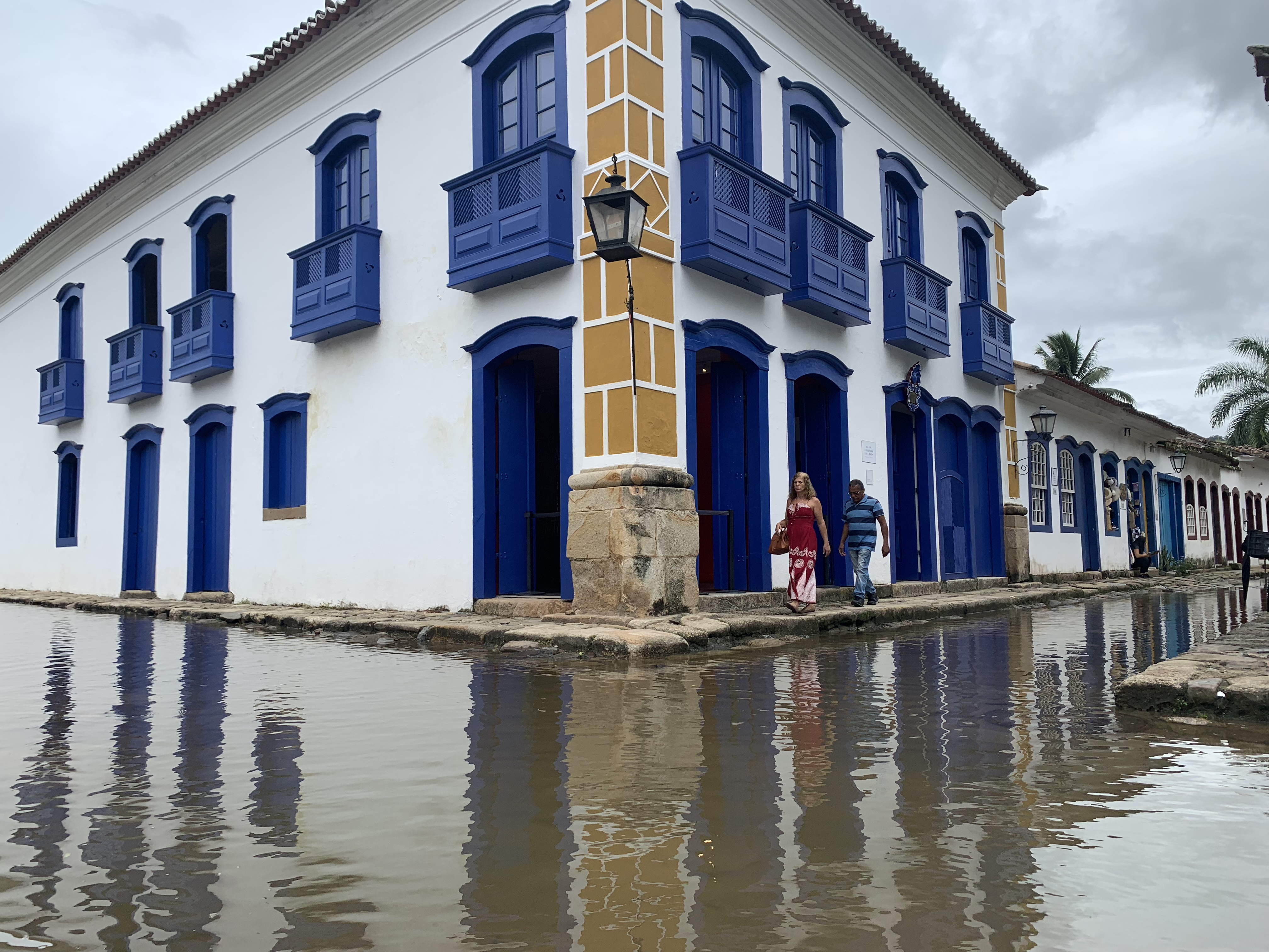 Duas pessoas vadejando pela alagada esquina de paralelepípedos em Paraty em frente ao icônico grande edifício de esquina em azul e ocre, sua fachada refletida na água da maré