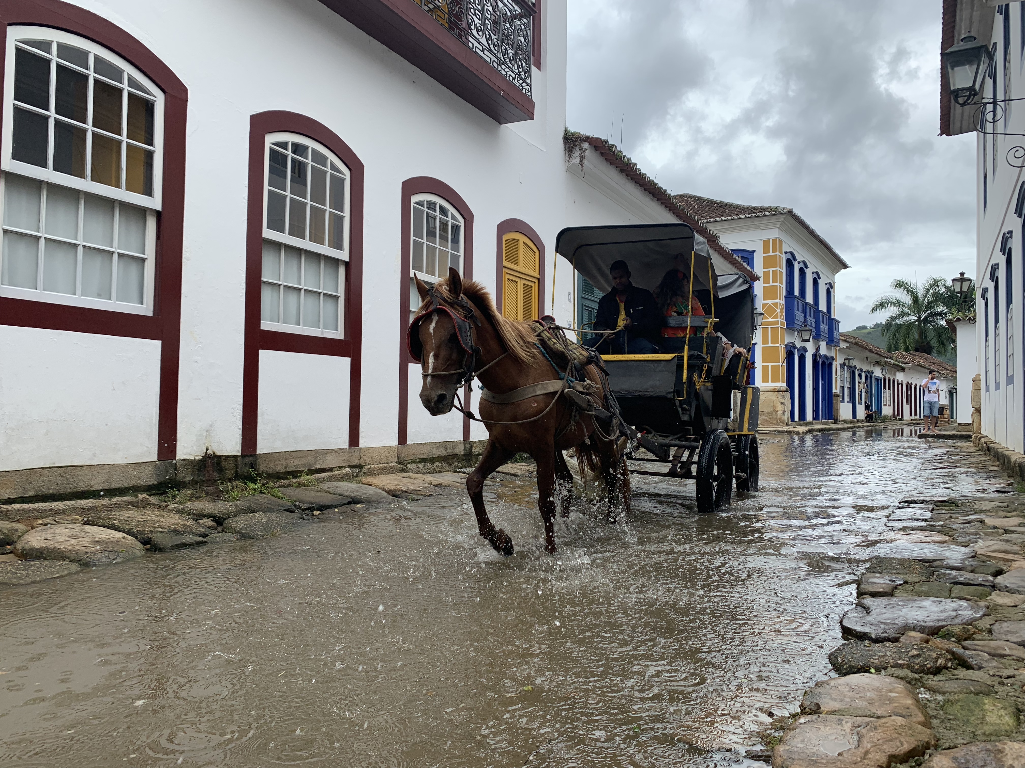 Uma charrete respingando a toda velocidade pela rua de paralelepípedos alagada em Paraty, os cascos do cavalo levantando grandes leques de spray, com edifícios coloniais dos dois lados