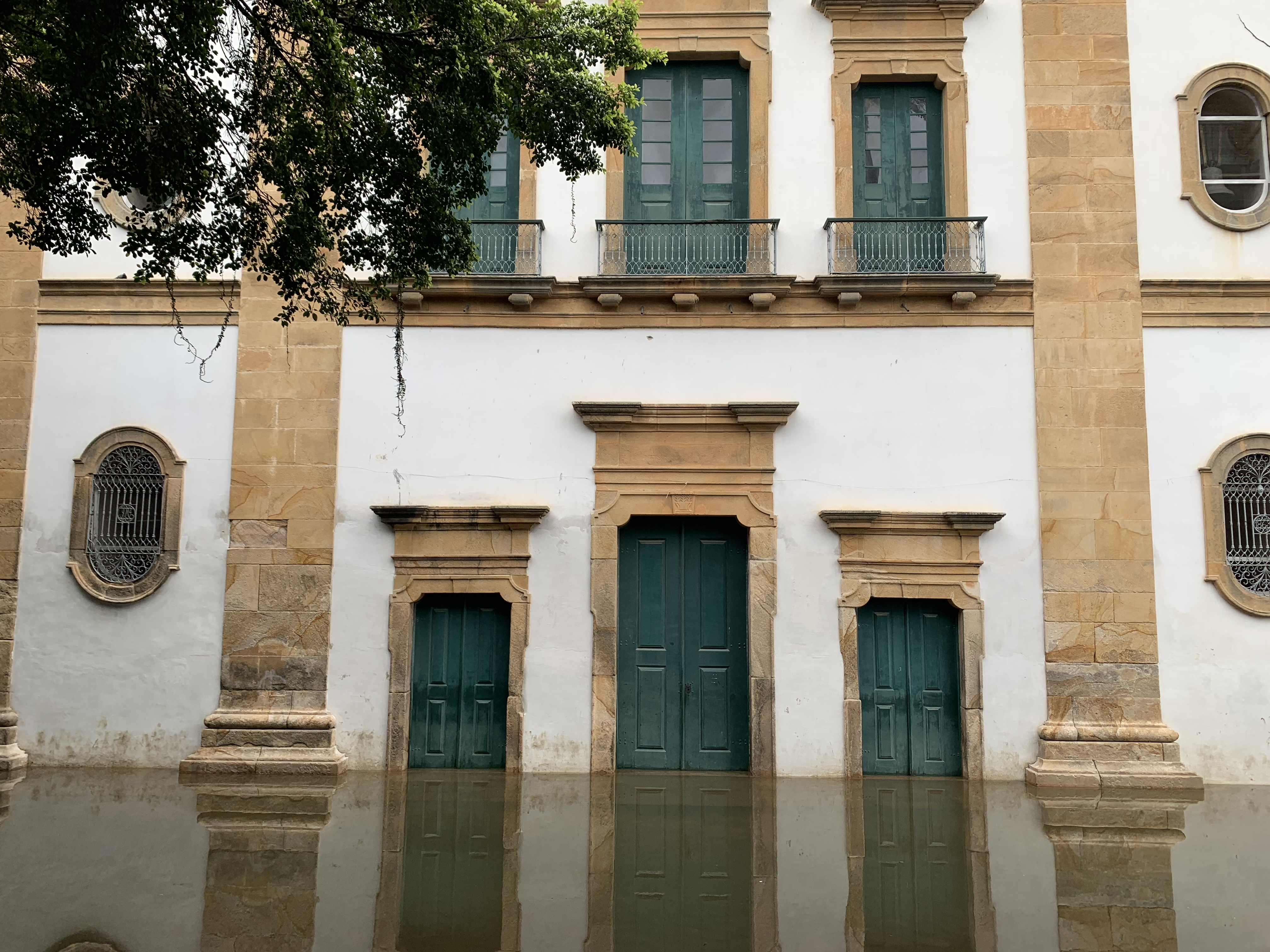 A fachada barroca de pedra de uma igreja colonial em Paraty completamente cercada pela enchente de maré em sua base, as portas e janelas com molduras de arenito erguendo-se acima de seus reflexos na água parada, um galho de árvore no canto superior esquerdo
