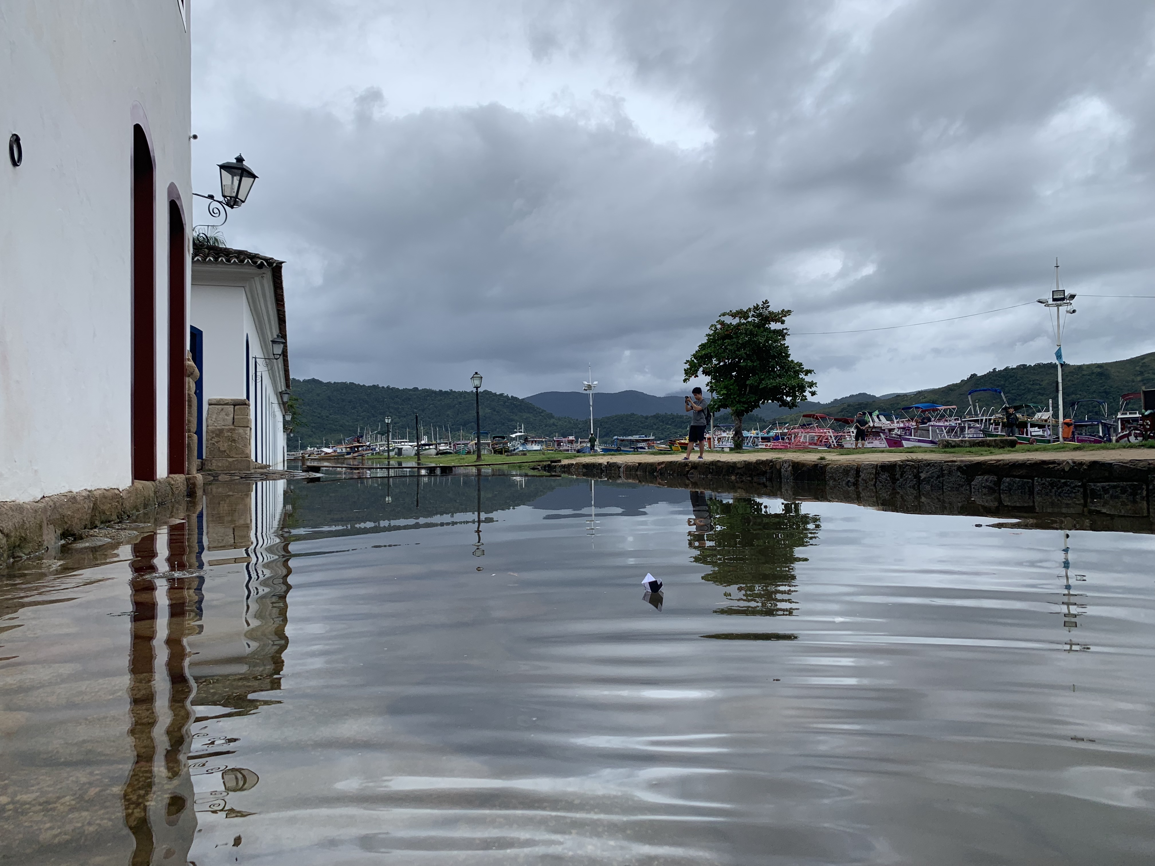O orla alagada de Paraty olhando para o calçadão completamente submerso em direção à marina e às montanhas verdes, um pequeno barco de papel flutuando em primeiro plano contra o céu dramático refletido na água parada