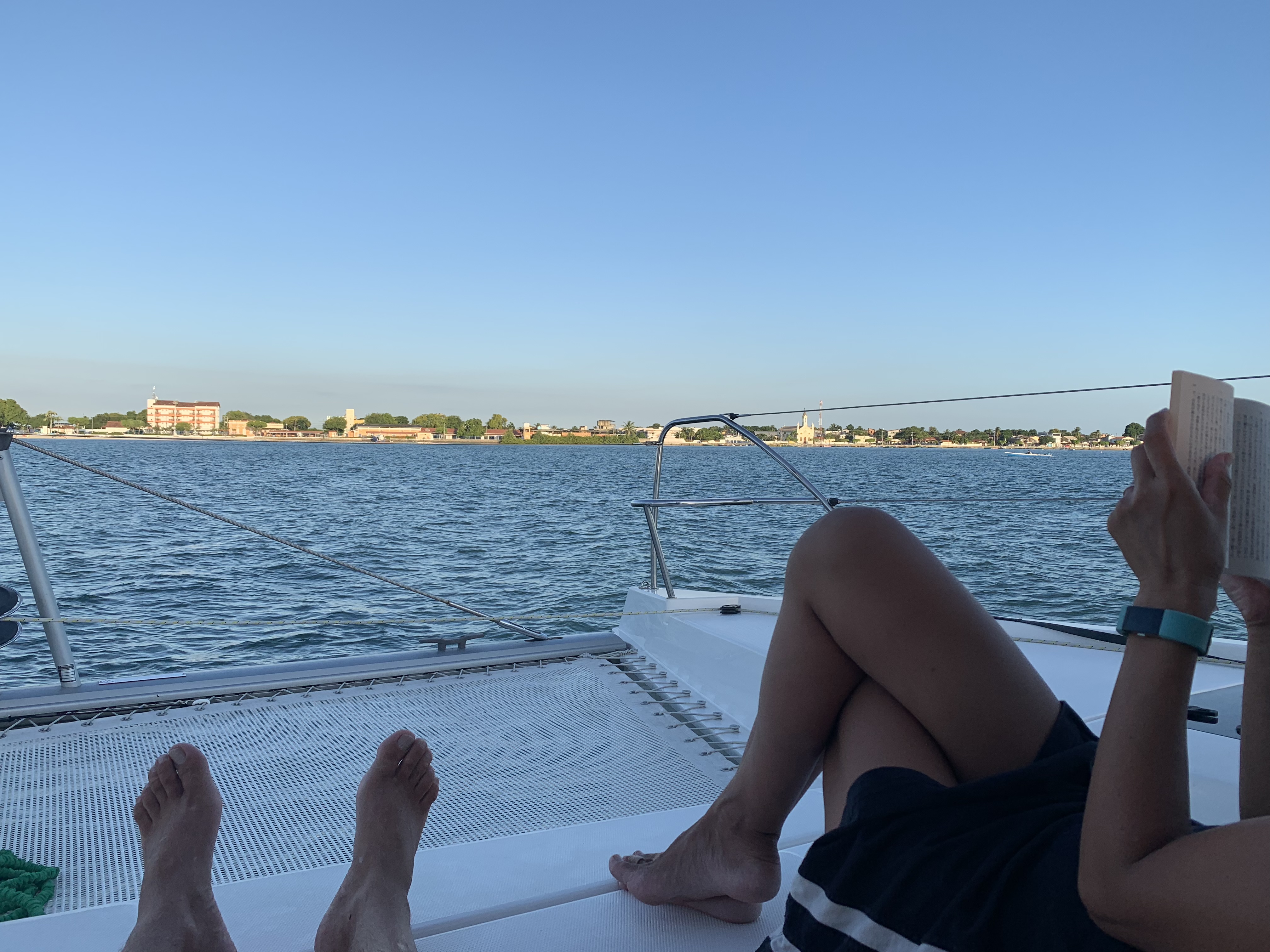 Yuka lendo no trampoline do catamarã fundeado em frente a Salinas da Margarida, com a cidade e o campanário da igreja visíveis através da água calma na luz dourada da tarde