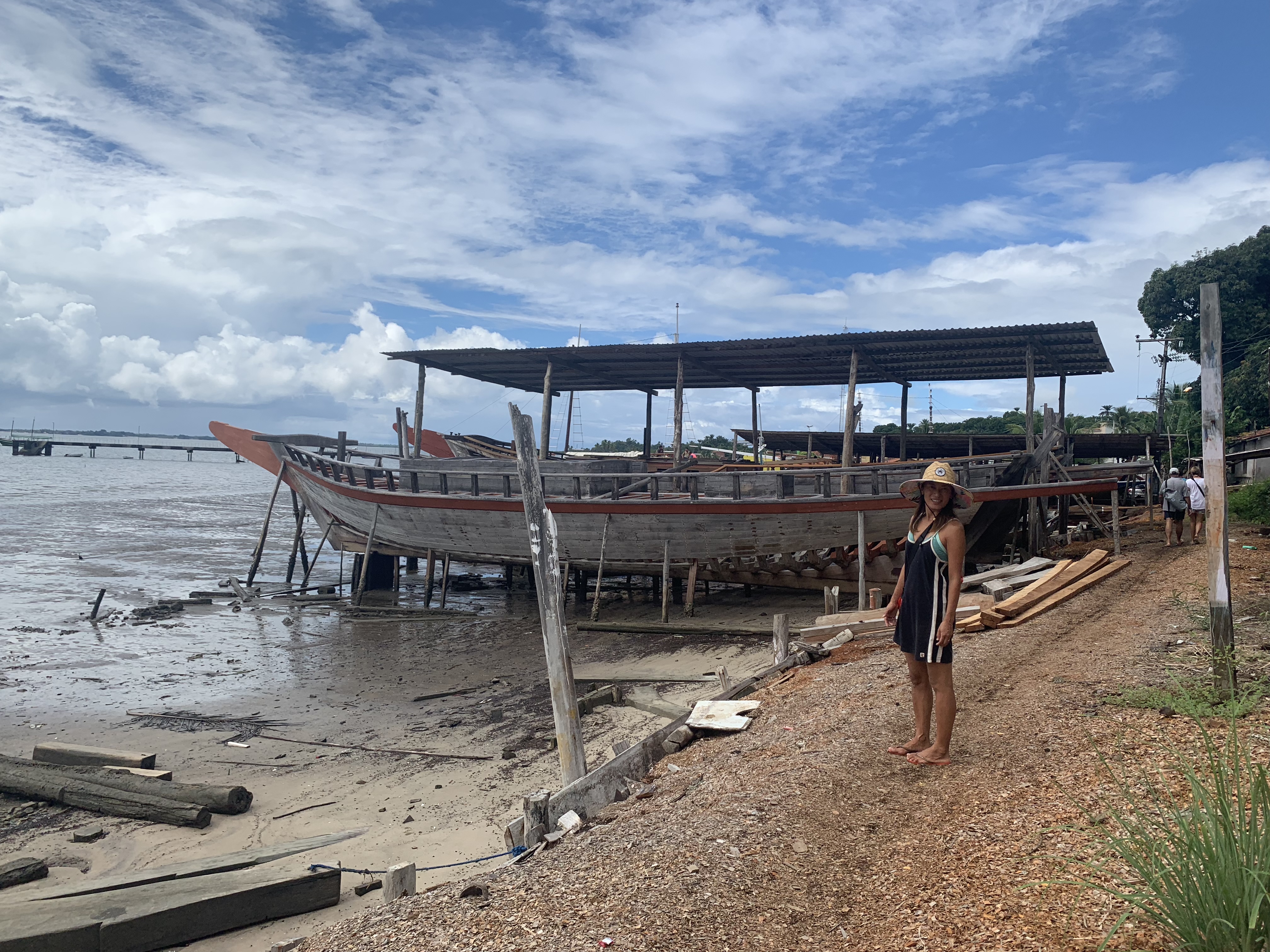 Yuka standing in front of a large wooden boat under construction at the Camamu boatyard